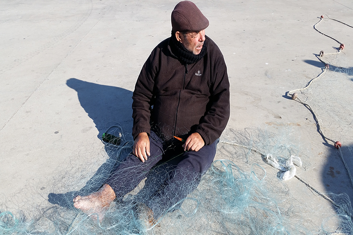 Remienda en el puerto de Adra 
La sombra de este pescador jubilado proyecta la tibia luz de una ma&ntilde;ana de invierno en el puerto de Adra (Almer&iacute;a). Sus saberes, su postura, sus pies y sus manos son las herramientas para rehacer el arte &ldquo;claro&rdquo;, llamado as&iacute; por su amplitud o claro de malla. La fisiolog&iacute;a y la cognici&oacute;n humana colaborando como artefactos t&eacute;cnicos clave en las pesquer&iacute;as artesanales. La flota de peque&ntilde;a escala, especializada en salidas hacia caladeros pr&oacute;ximos y organizada sobre relaciones familiares mayoritariamente, se ha convertido en un sector refugio en muchos puertos pesqueros andaluces. Los marineros de m&aacute;s edad siguen acometiendo tareas insustituibles por su pericia t&eacute;cnica y su resistencia f&iacute;sica y ps&iacute;quica.